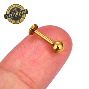 May include: Gold titanium body jewelry with a ball end. The jewelry is on a finger and the text "IMPLANT GRADE TITANIUM G23 ASTM F136" is on a white circle.