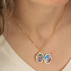May include: A gold chain necklace with a locket pendant. The locket is open and shows two photos of dogs inside. The locket has a scalloped edge.