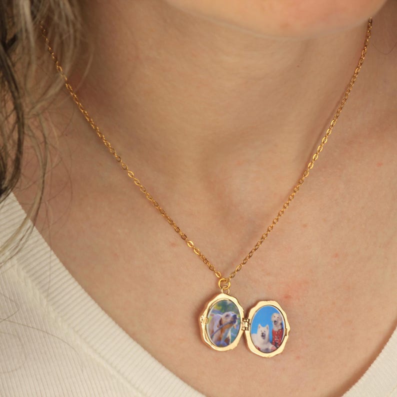 May include: A gold chain necklace with a locket pendant. The locket is open and shows two photos of dogs inside. The locket has a scalloped edge.