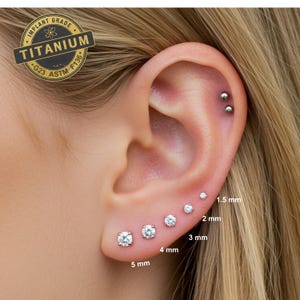 May include: Close-up of a person's ear with five different sizes of clear round stud earrings. The earrings are labeled with their size in millimeters: 1.5 mm, 2 mm, 3 mm, 4 mm, and 5 mm.