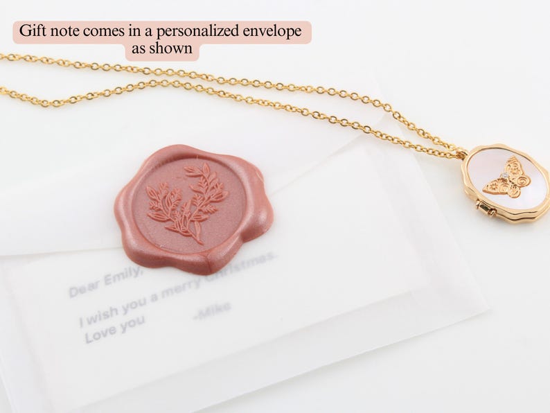 May include: A gold chain necklace with a white and gold locket featuring a butterfly design. The necklace is packaged in a white envelope with a red wax seal.
