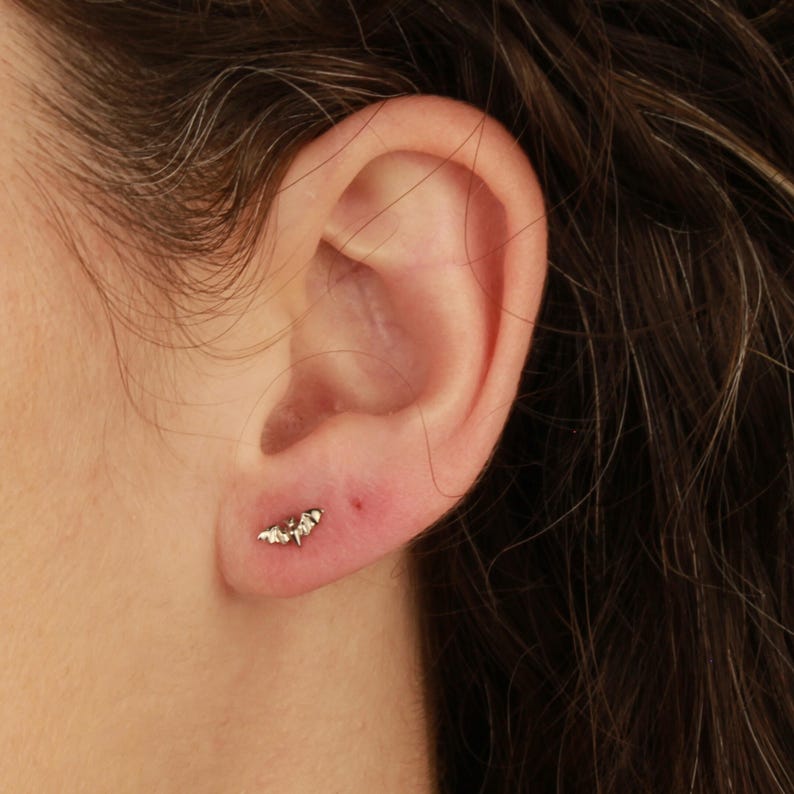 K&ouml;nnte beinhalten: Nahaufnahme eines Ohrs mit einem kleinen, silbernen Ohrring in Form einer Fledermaus. Der Ohrring befindet sich im Ohrl&auml;ppchen.