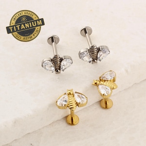 May include: A collection of bee-shaped earrings. Two are silver-toned with clear, teardrop-shaped gems for wings. Two are gold-toned with similar gems. The text "IMPLANT GRADE TITANIUM G23 ASTM F136" is in the upper left corner.
