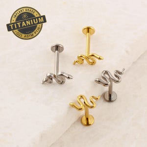May include: Three surgical steel body jewelry pieces with a snake design. The snake is gold colored and the posts are silver. The jewelry is labeled as implant grade titanium G23 ASTM F136.