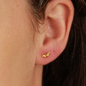 K&ouml;nnte beinhalten: Nahaufnahme eines Ohres mit einem kleinen goldenen Fledermaus-Ohrring. Der Ohrring ist ein schlichtes Design mit einer zarten Fledermaus-Silhouette.