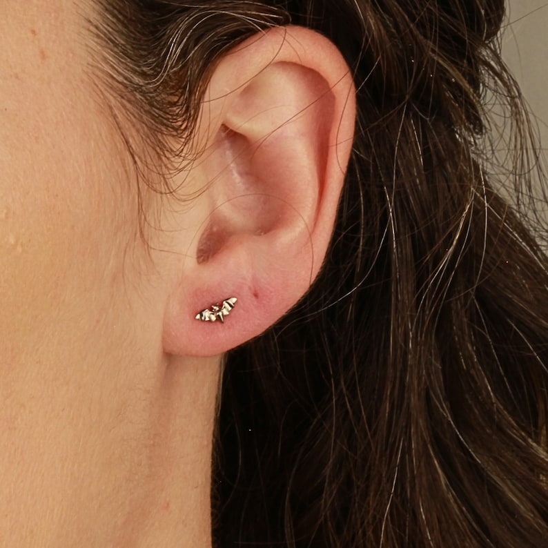 K&ouml;nnte beinhalten: Nahaufnahme eines Ohres mit einem kleinen silbernen Fledermaus-Ohrring. Der Ohrring ist ein schlichtes Design mit einer zarten Fledermausform.