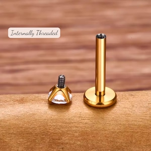K&ouml;nnte beinhalten: Innengewindetes Gold-K&ouml;rperpiercing mit einem klaren Edelstein-Oberteil. Der Schmuck liegt auf einer Holzoberfl&auml;che.