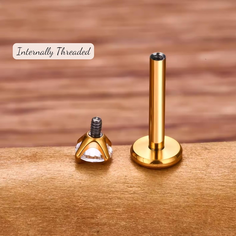 K&ouml;nnte beinhalten: Innengewindetes Gold-K&ouml;rperpiercing mit einem klaren Edelstein-Oberteil. Der Schmuck liegt auf einer Holzoberfl&auml;che.