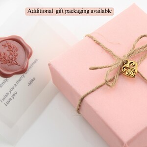 May include: A pink gift box tied with twine and a gold heart charm. The box is sitting on a white surface with a clear plastic bag with a red wax seal and a handwritten note that says "Dear Emily, I wish you a merry Christmas. -Mike". The text "Additional gift packaging available" is written in black text on a white background.