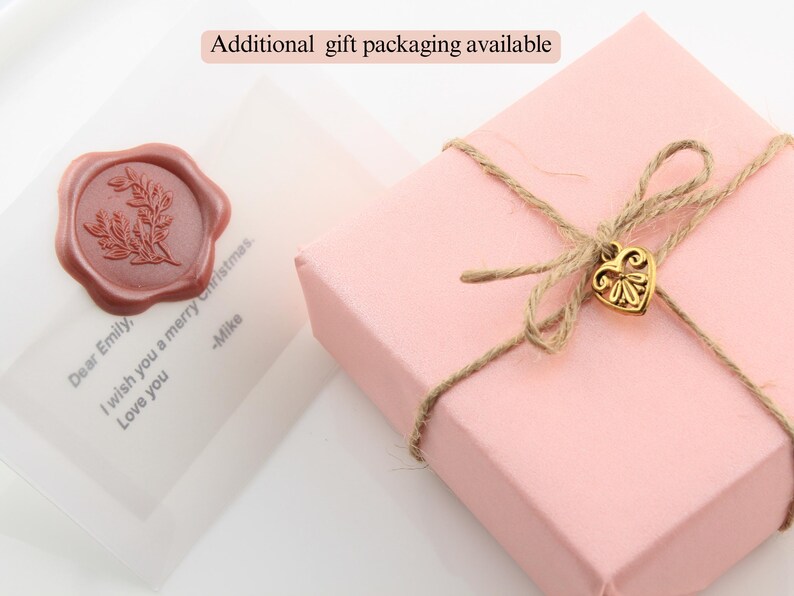 May include: A pink gift box tied with twine and a gold heart charm. The box is sitting on a white surface with a clear plastic bag with a red wax seal and a handwritten note that says "Dear Emily, I wish you a merry Christmas. -Mike". The text "Additional gift packaging available" is written in black text on a white background.