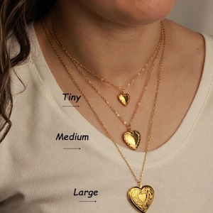 May include: Three gold-colored heart-shaped necklaces of varying sizes: tiny, medium, and large. The necklaces are layered, with the smallest heart pendant at the top and the largest at the bottom. The word "Tiny", "Medium", and "Large" are written on the image.
