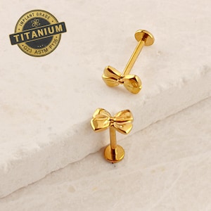 May include: Two gold bow-shaped body jewelry pieces with a post and a ball. The jewelry is made of titanium and is implant grade.