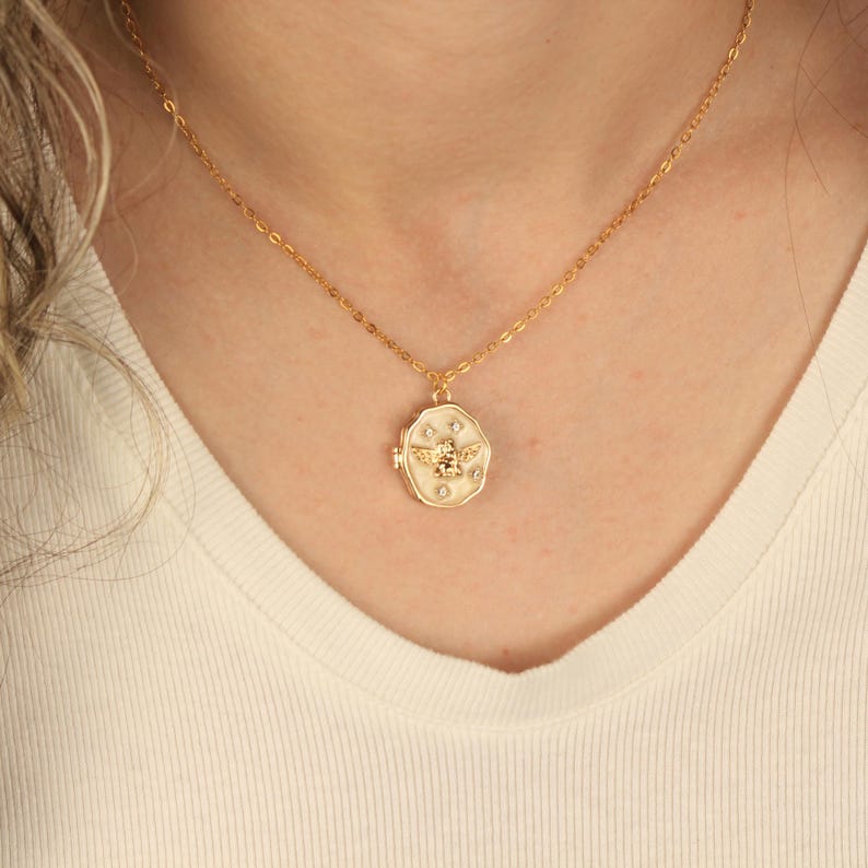 May include: A gold chain necklace with a white enamel locket pendant featuring a bee design and small clear rhinestones.