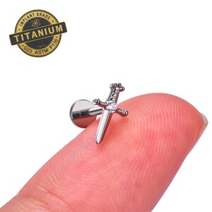 May include: A silver-colored sword-shaped earring with a clear gemstone in the center. The earring is made of titanium and has a flat back. The image also includes a gold-colored stamp that reads "IMPLANT GRADE TITANIUM G23 ASTM F136."