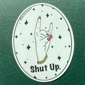 May include: White circular sticker with a black outline. A black and white illustration of a hand giving the middle finger with red nail polish. The hand is wearing a gold chain bracelet. The text "Shut Up." is printed in black below the hand. The background is white with black stars.