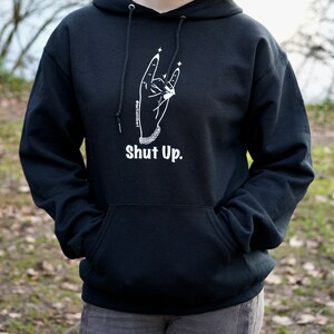 May include: Black hoodie with a white graphic of a hand making the rock and roll hand gesture with the text "Shut Up." printed below.