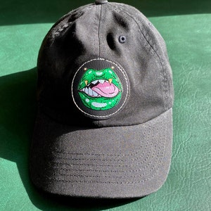 Puede incluir: Una gorra de béisbol negra con un parche de boca verde y dentada. El parche tiene una lengua rosa y dientes blancos.