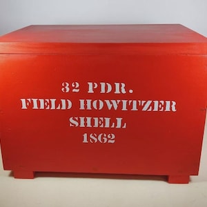 May include: A red wooden box with a hinged lid and side handles. The box has white lettering that reads "32 PDR. FIELD HOWITZER SHELL 1862". The box is rectangular and sits on four small feet.