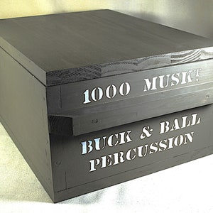 Puede incluir: Una caja de madera gris oscuro con tapa con bisagras. La caja tiene letras blancas que dicen "1000 MUSK!" y "BUCK & BALL PERCUSSION". La caja es rectangular y parece hecha a mano.