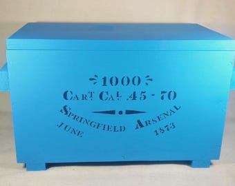 May include: A light blue wooden box with a lid and handles. The front of the box features stenciled text that reads "1000 CART CAL .45-70 SPRINGFIELD ARSENAL JUNE 1873".