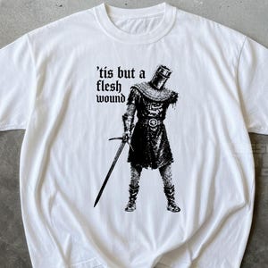 May include: A white short-sleeved t-shirt featuring a black graphic illustration of a medieval knight missing an arm, holding a sword. The text 'tis but a flesh wound' is printed in a black gothic-style font to the left of the knight figure.