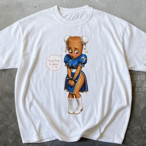 Könnte beinhalten: Ein weißes Kurzarm-T-Shirt mit einer zentralen Grafik einer braunhäutigen Figur mit Haarknoten, die ein blaues Kleid, weiße Stiefel und Stachelarmbänder trägt. Eine Sprechblase neben der Figur enthält den Text Tung Tung... S-so...