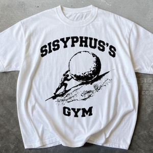 Könnte beinhalten: Ein weißes kurzärmeliges T-Shirt mit einem schwarzen Grafikdruck auf der Brust. Das Design zeigt eine Figur, die einen großen Felsbrocken einen Hügel hinaufschiebt, mit dem Text "SISYPHUS'S" darüber und dem Wort "GYM" darunter gedruckt.