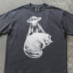 May include: A charcoal grey short-sleeve t-shirt featuring a white graphic print of a cat being lifted by a flying saucer. The design includes a beam of light extending from the UFO down to the cat, with the text DESIGN TEES CENTRAL visible.