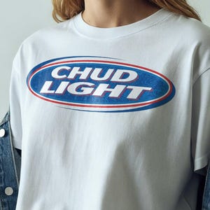 Può includere: T-shirt bianco con la scritta "CHUD LIGHT" in un design ovale blu e rosso. La maglietta è indossata con una giacca di jeans e pantaloni grigi. Uno stile di abbigliamento casual.