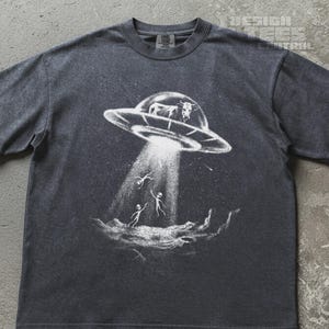 May include: A charcoal grey short-sleeve t-shirt featuring a white graphic print of a flying saucer abducting a cow. The design shows a beam of light extending from the UFO towards the ground, with two small alien figures standing below.