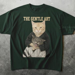 Puede incluir: Una camiseta verde oscuro con un gráfico de un gato naranja vestido con un gi de artes marciales negro, sosteniendo a un gato gris más pequeño que tiene la lengua fuera. Sobre los gatos, el texto THE GENTLE ART aparece en fuente beige.