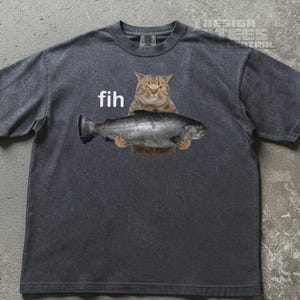 Op de afbeelding: Een antracietgrijs T-shirt met korte mouwen en een grafische print van een bruine cyperse kat die een grote zilveren vis vasthoudt. De tekst "fih" staat in witte kleine letters boven de vis gedrukt. Het shirt heeft een ronde halslijn.