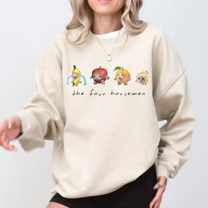 Puede incluir: Una sudadera beige con un gráfico de cuatro gatos vestidos de fruta. Los gatos están vestidos de plátano, manzana, naranja y melocotón. El texto "the four horsemen" está impreso debajo del gráfico.