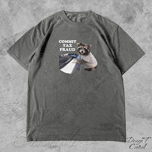 May include: A dark grey t-shirt featuring a graphic of a raccoon wearing a tie and a white shirt, sitting at a computer keyboard. The text "COMMIT TAX FRAUD" is above the raccoon.