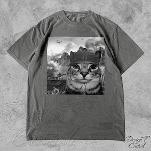 Peut inclure: Un t-shirt gris avec une image en noir et blanc d'un chat portant un casque militaire. Le chat regarde directement le spectateur. L'image est sur fond de scène de guerre avec des montagnes, des chars et des avions.