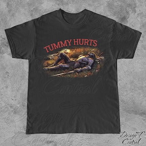 May include: Black t-shirt featuring a red graphic of a knight lying down in a field with the text "TUMMY HURTS" above the image.