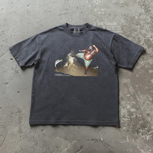May include: Dark gray t-shirt featuring a graphic of a person in camouflage shorts kicking. The graphic is set against a brown and tan background. The shirt has short sleeves and a crew neck.