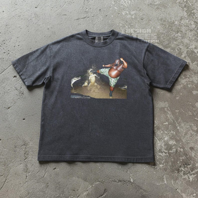 May include: Dark gray t-shirt featuring a graphic of a person in camouflage shorts kicking. The graphic is set against a brown and tan background. The shirt has short sleeves and a crew neck.