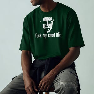 May include: Dark green t-shirt with a white graphic of a face wearing glasses and the text "fuck my chud life". The shirt is made of a soft material and has short sleeves. The person is wearing gray pants.