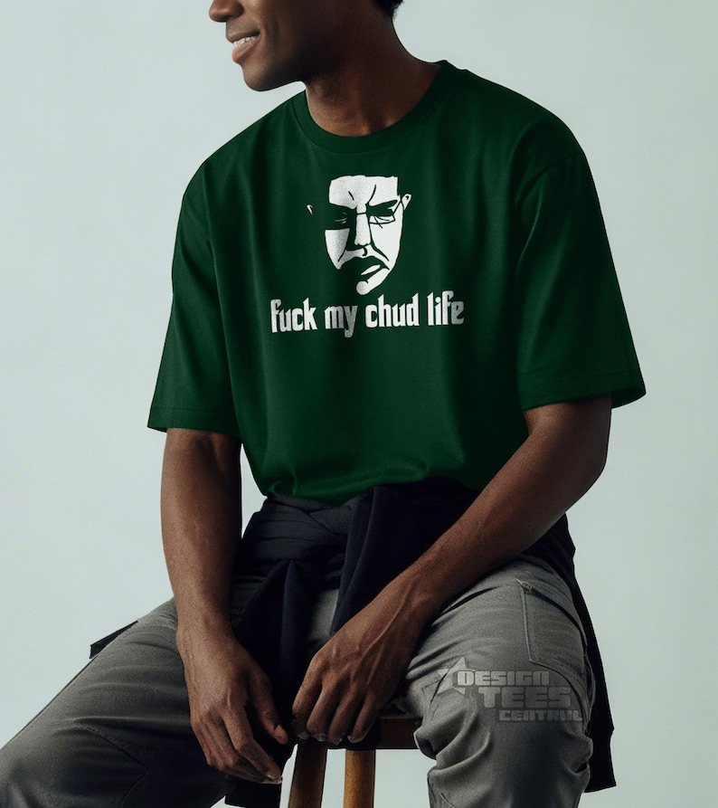 May include: Dark green t-shirt with a white graphic of a face wearing glasses and the text "fuck my chud life". The shirt is made of a soft material and has short sleeves. The person is wearing gray pants.