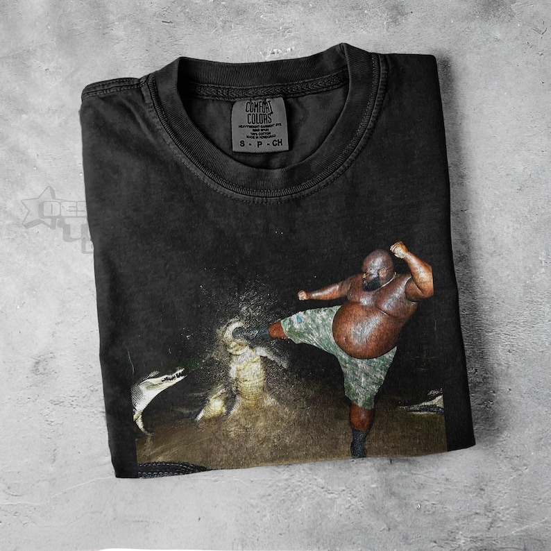 May include: Black Comfort Colors t-shirt featuring a graphic of a person kicking a snake. The person is wearing camouflage shorts. The shirt has a tag that says Comfort Colors.