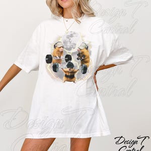 May include: White t-shirt featuring a whimsical design of three hamsters lifting weights under a full moon. The hamsters wear hats and headphones, with a lightning bolt effect around them. The shirt has a relaxed fit.