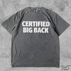 May include: Grey t-shirt featuring the white text "CERTIFIED BIG BACK". The shirt has a crew neck and short sleeves. The fabric appears to be a soft, washed material. A casual, everyday garment.