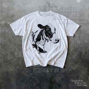 May include: White t-shirt with a black and white illustration of a cow on a skateboard. The cow has black markings and a halter. The shirt is made of a soft material and has a crew neck.