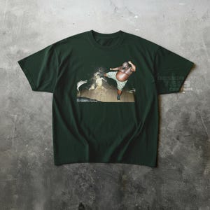 May include: Dark green t-shirt featuring a graphic of a person kicking a creature in water. The person is wearing camouflage shorts. The text "DESIGN TEES CENTRAL" is visible on the right side of the shirt.