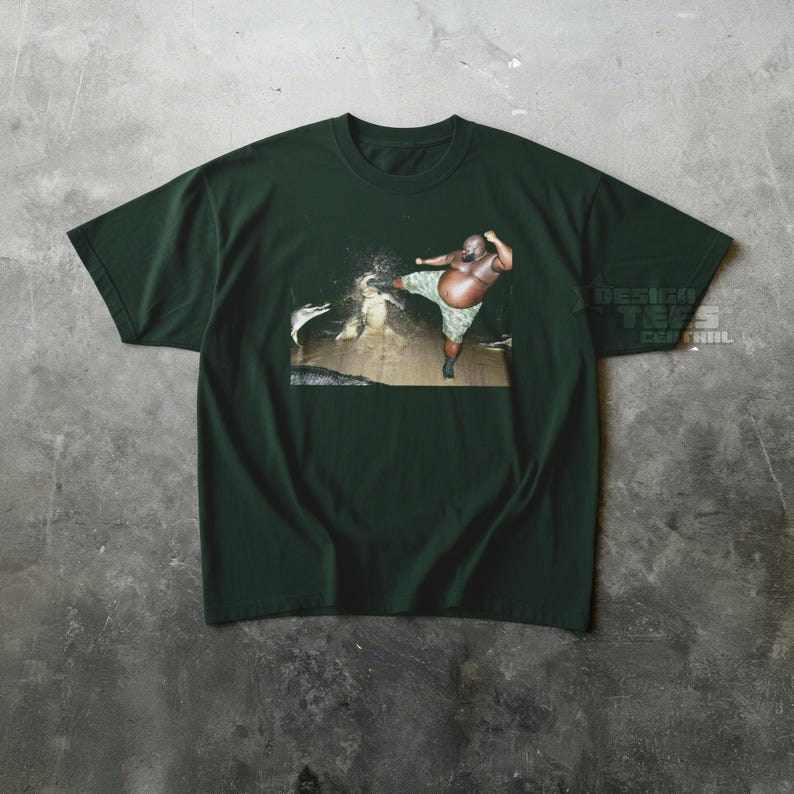 May include: Dark green t-shirt featuring a graphic of a person kicking a creature in water. The person is wearing camouflage shorts. The text "DESIGN TEES CENTRAL" is visible on the right side of the shirt.