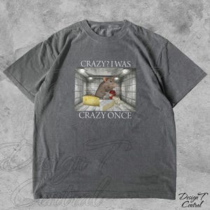 Puede incluir: Camiseta gris con un gr&aacute;fico de una rata en una jaula, queso y el texto "Crazy? I was crazy once". La rata lleva un guante de boxeo rojo. La camiseta tiene un corte holgado y est&aacute; hecha de material suave.