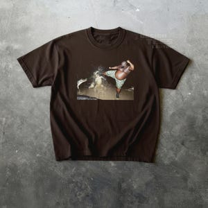 May include: Brown t-shirt featuring a graphic of a person kicking an alligator. The person is wearing camouflage shorts. The image is set against a dark background. The shirt has a crew neck and short sleeves.
