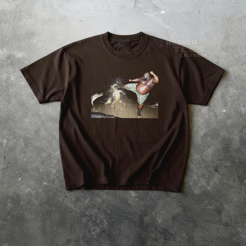 May include: Brown t-shirt featuring a graphic of a person kicking an alligator. The person is wearing camouflage shorts. The image is set against a dark background. The shirt has a crew neck and short sleeves.
