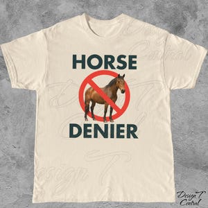 Peut inclure: T-shirt cr&egrave;me avec l'inscription "HORSE DENIER" en bleu fonc&eacute;. Un cheval brun est repr&eacute;sent&eacute; &agrave; l'int&eacute;rieur d'un cercle rouge barr&eacute;. Le t-shirt a un col rond et des manches courtes.
